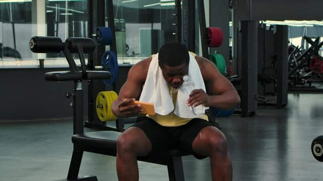 African American man looks at phone in gym, drinks water while relaxing, then celebrates victory received good news. Sports facilities in background for fitness and bodybuilding. Close up. Slow motion