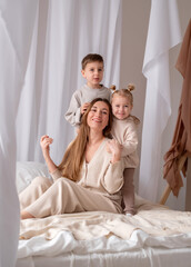 Obraz na płótnie Canvas Portrait of smiling young mommy with two kids sitting on the bed. Attractive mother and two children, son and daughter are laughs in bedroom.