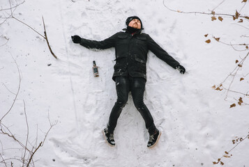 A severely drunk homeless man in a black coat and hat lies, asleep on his back on white snow in the...