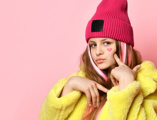 Close up portrait of modern bright confident teen girl on pink background. Child with pink strands of hair dressed in a yellow coat and hat points to a painted heart on his face. Place for text.