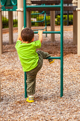 A white toddler boy is playing at a playground alone. He is holding the bars of a metal ladder and trying to pull himself up as he learns to climb. Surface is covered with wood chips for added safety