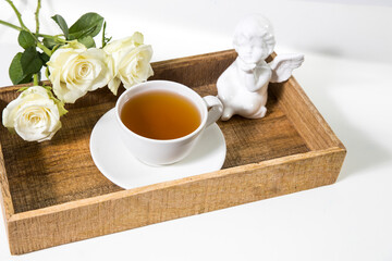 A bouquet of white roses in a vintage glass vase, a wooden tray with a cup of tea, a faience figurine of an angel. Greeting card for mother's day.