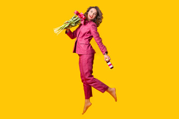Fototapeta na wymiar Photo of happy woman in suit jumping over yellow background and holding bunch of tulips and fresh bottle juice.