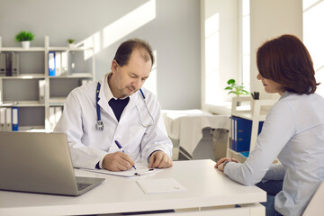 Healthcare in a hospital or private clinic. Middle-aged doctor writes a prescription to a female patient in his hospital office. Concept of disease prevention and consultations in the medical clinic.