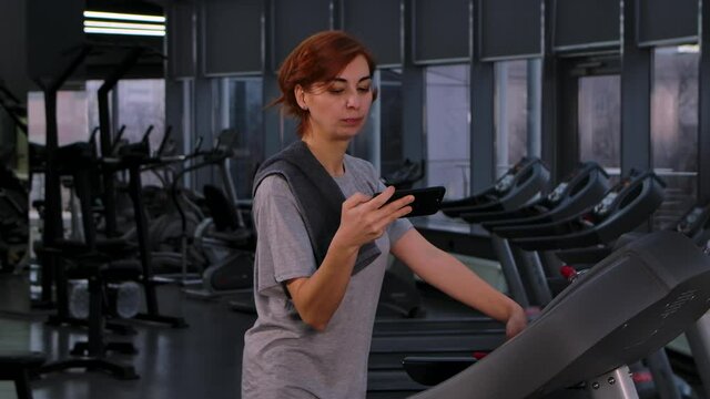 Young woman doing speedwalk on treadmill looks at smartphone, then shocked and excited. Surprised joyful female in sportswear on background of gym rejoices at good news. Close up. Slow motion.