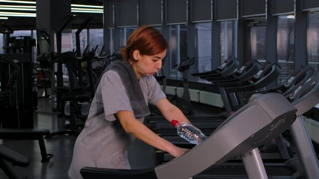 Tired sweaty young woman runs on a treadmill in the gym. Athlete female in sportswear finishes cardio workout and towels off. Sports equipment in the background for fitness. Close up. Slow motion.
