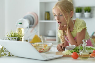 Obraz na płótnie Canvas cute girl making salad on kitchen and using laptop