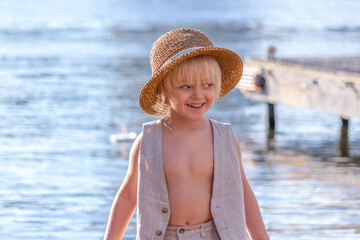 Happy little blond caucasian boy wearing straw hat stands by the river. Summer kids fun theme.