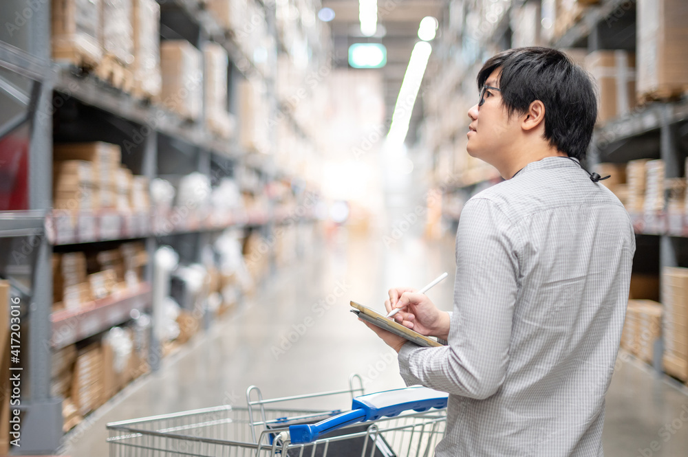 Wall mural Asian man worker doing stocktaking of product in cardboard box on shelves in warehouse by using digital tablet and pen. Physical inventory count concept