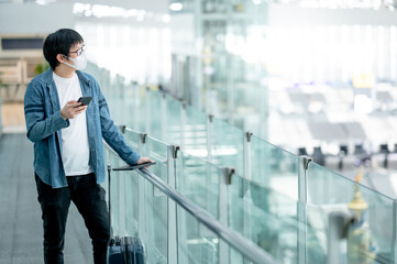 Asian man tourist with suitcase luggage wearing face mask using smartphone in airport terminal. Coronavirus (COVID-19) pandemic prevention when travel abroad. Health awareness and social distancing
