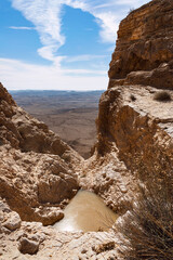 rainpool in a narrow canyon at the rim of the makhtesh ramon crater in israel with the bottom of the crater and a partly cloudy blue sky in the background