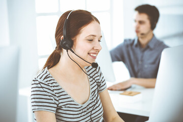 Casual dressed young woman using headset and computer while talking with customers online. Group of operators at work. Call center, business concept