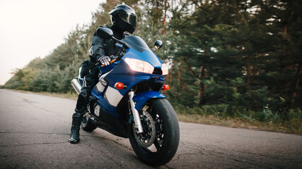 Motorcyclist in leather protective suit and black helmet sits on sports motorcycle. Biker in black rides on the road against the background of the forest