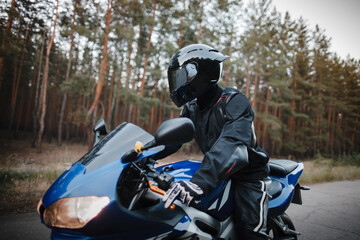 Motorcyclist in leather protective suit and black helmet sits on sports motorcycle. Biker in black rides on the road against the background of the forest