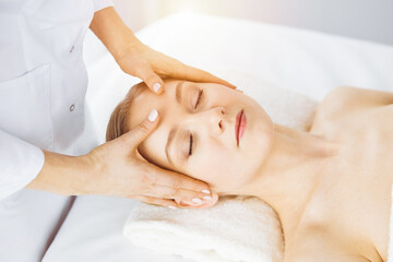 Obraz na płótnie Canvas Beautiful caucasian woman enjoying facial massage with closed eyes in sunny spa salon. Relaxing treatment in medicine