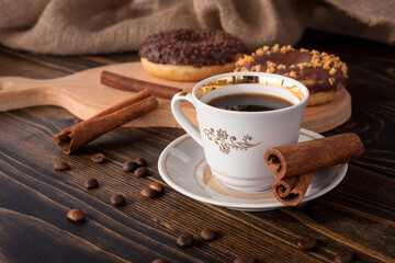 A cup of aromatic black coffee and coffee beans on the table with cinnamon sticks. Morning espresso coffee for breakfast in a beautiful white cup. With a delicious donut in the background.
