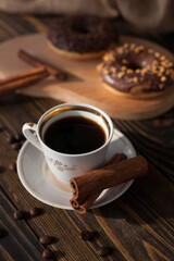 A cup of aromatic black coffee and coffee beans on the table with cinnamon sticks. Morning espresso coffee for breakfast in a beautiful white cup. With a delicious donut in the background.