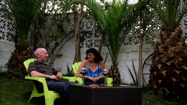 Senior couple drinking wine, talking and relaxing in the garden under palm trees in the evening.