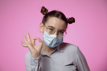Modern young teen girl in protective medical facial mask showing okay gesture on pink studio background with copy space