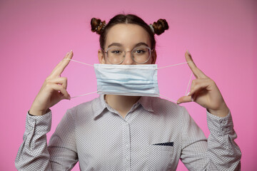 Modern stylish young teen girl putting on medical mask protecting from coronavirus pandemic on pink studio backdrop