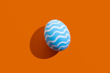 Easter art. Festive ornament. Spring holiday. Handmade decoration. Abstract background. Blue egg with white pattern waves isolated on orange copy space.