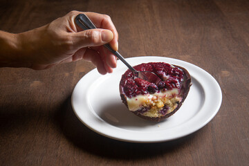 Hand puts a spoon in an opened and cracked red fruits stuffed easter egg on a white plate on a wooden table.