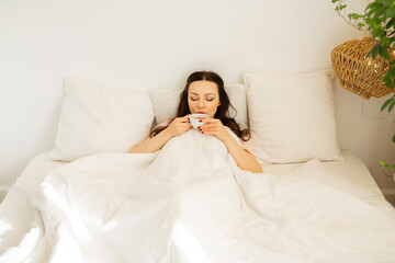 beautiful young woman lies on the bed under the covers with a cup of tea 
