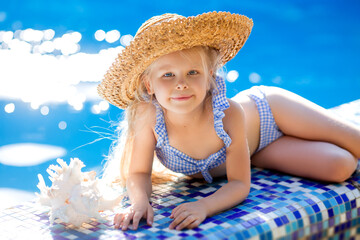 little blonde girl in a striped swimsuit and a straw hat is lying on the side of the pool next to a...