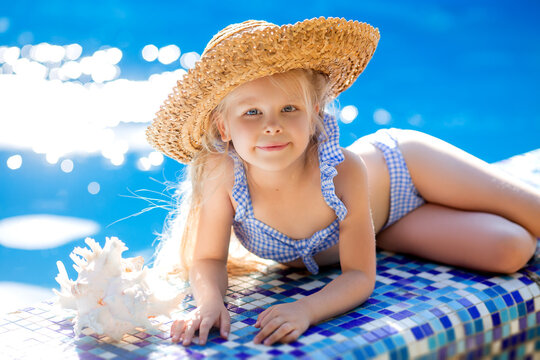 little blonde girl in a striped swimsuit and a straw hat is lying on the side of the pool next to a huge white shell