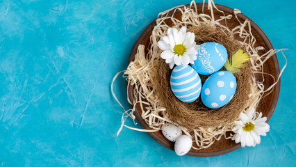 Natural decoration. Happy Easter. Holiday composition. Organic painted eggs with creative pattern white flowers feather in nest in rustic plate with straw on blue textured copy space background.