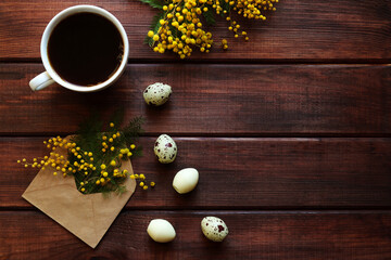 Easter greeting card. Rustic spring texture with mimosa flowers, quail eggs-shaped sweets and cup of coffee on dark wooden background with copy space. Flat lay, top view
