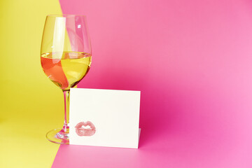 A glass of white wine on a pink and yellow background, card with a lip print with lipstick, the concept of the on March 8