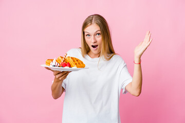 Young russian woman eating a waffle isolated receiving a pleasant surprise, excited and raising hands.