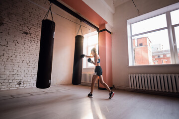 Female kickboxer hitting punching bag while dust particles flies in sunflare light background.