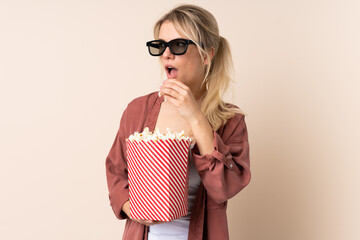 Blonde woman over isolated background with 3d glasses and holding a big bucket of popcorns while looking side