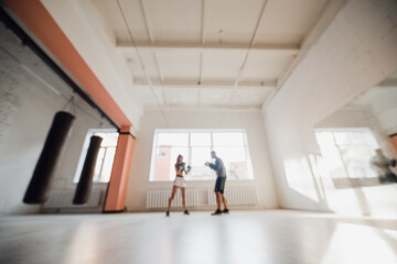 Fototapeta premium Medium shot of young woman doing boxing workout at the gym and punching her instructor, who is dodging her blows and controlling training process.