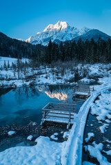 romantic path through snow along a beautiful emerald, clear volcanic lake in winter at a touristic nature reserve in the Alps. Reflection of snowcapped mountains glowing in the rising sun. no people