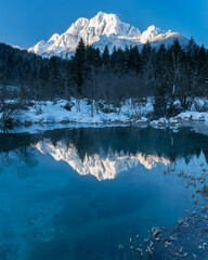 romantic path through snow along a beautiful emerald, clear volcanic lake in winter at a touristic nature reserve in the Alps. Reflection of snowcapped mountains glowing in the rising sun. no people