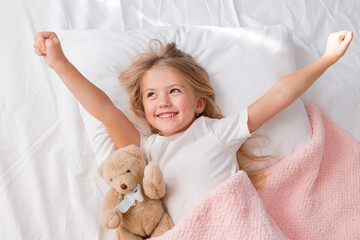 little blonde girl sleeping in bed hugging a teddy bear under a pink knitted blanket, space for text