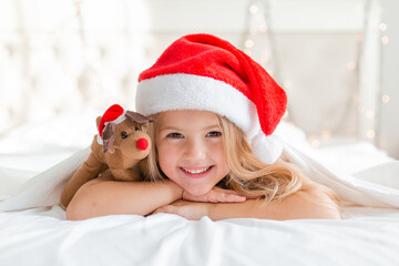 little blonde girl in a red Santa Claus hat is lying on her stomach under a blanket with a toy deer