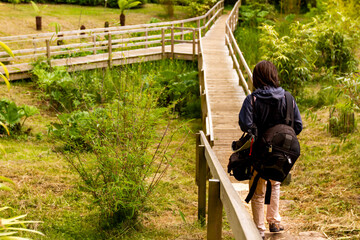 a young adventurer woman walking alone on a narrow wooden hiking path carrying big bags. The path bifurcates ahead at which point she needs to make a decision. Abstract concept image.