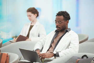 Naklejka na ściany i meble Portrait of modern African American man wearing lab coat while using laptop at coworking center in medical college, copy space