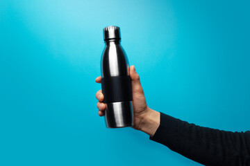 Close-up of male hand holding reusable steel water bottle against blue background.