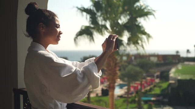 Attractive young woman in bathrobe taking pictures of gorgeous tropical landscape while enjoying the view, standing on hotel balcony. Vacation, technology concept