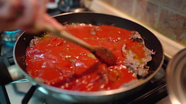 Person stirring meat slices in tomato sauce with wooden spoon, chef cooking traditional italian pizzaiola dish at home. Veal fillets frying in tomato sauce with different herb spices