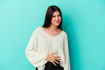 Fototapeta premium Young caucasian woman isolated on blue background touches tummy, smiles gently, eating and satisfaction concept.