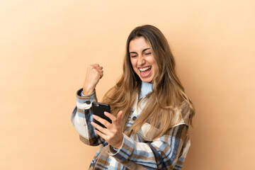 Young caucasian woman isolated on beige background with phone in victory position