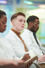 Vertical portrait of young man wearing lab coat while sitting in row in audience and listening to...