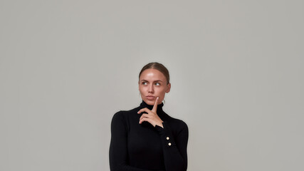 Portrait of dreamy caucasian young woman wearing black turtleneck holding finger near her face, looking thoughful aside, standing isolated over gray background