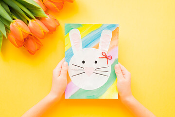 Kid's hands holding hand-made Easter card and orange tulips on yellow background. Top view. Easter concept. 